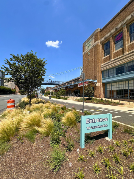 View north along Columbus Boulevard showing Pier 5 building, Ben Franklin Bridge in background, and teal “Entrance 7 N Columbus Blvd” sign in landscaped median.