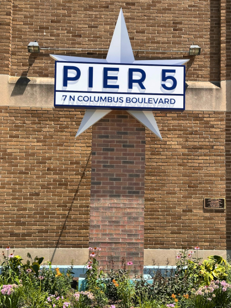 Close-up of white star sign reading “PIER 5 — 7 N Columbus Boulevard” mounted on brick wall above flower bed.